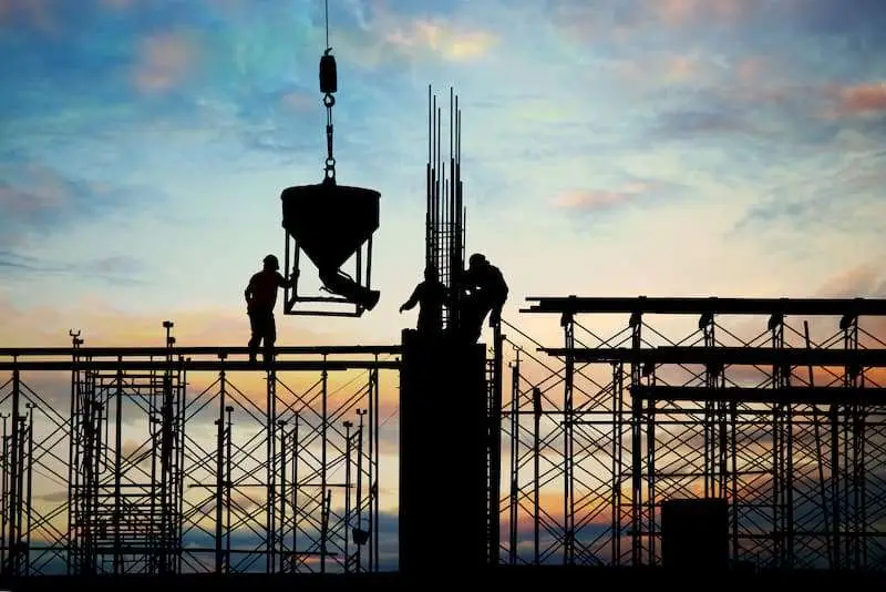 The silhouette of construction workers on a scaffolding against a colorful sunset sky, with cranes and structures in the background.