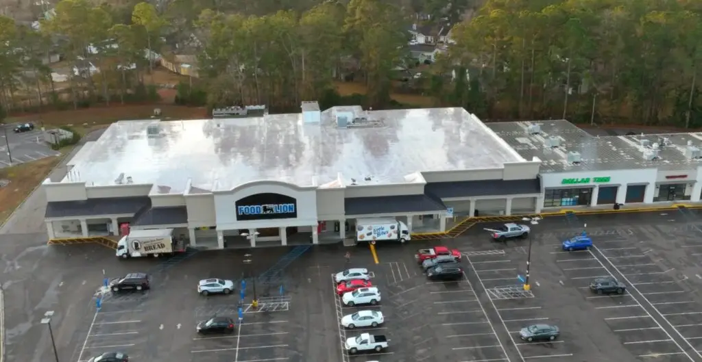 An aerial view of a shopping plaza with a large grocery store and various other retail businesses, surrounded by a paved parking lot and trees